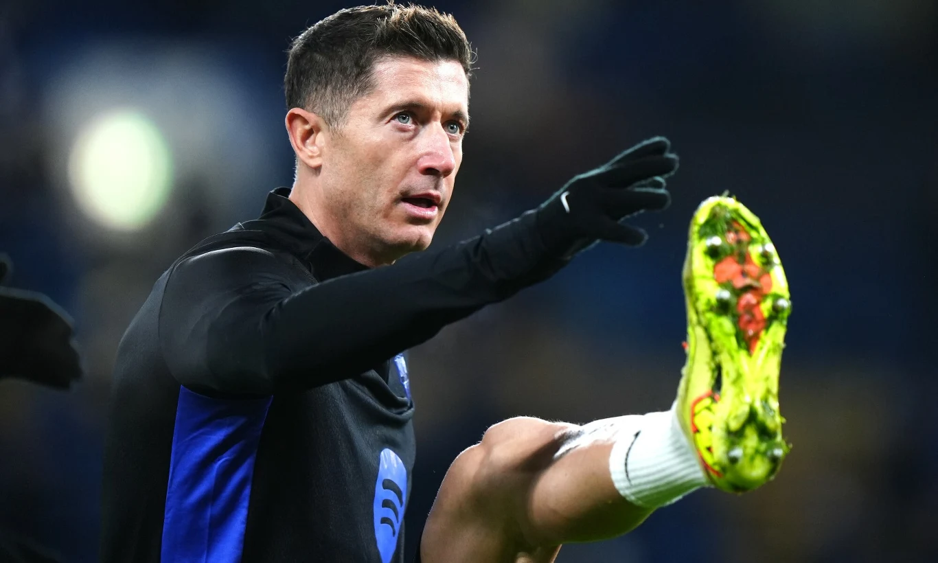 Robert Lewandowski Mężczyzna w sportowym stroju wykonuje rozgrzewkowe ćwiczenie, unosi nogę wysoko do góry, a na pierwszym planie widoczny jest jasny, neonowy but piłkarski pokryty trawą.