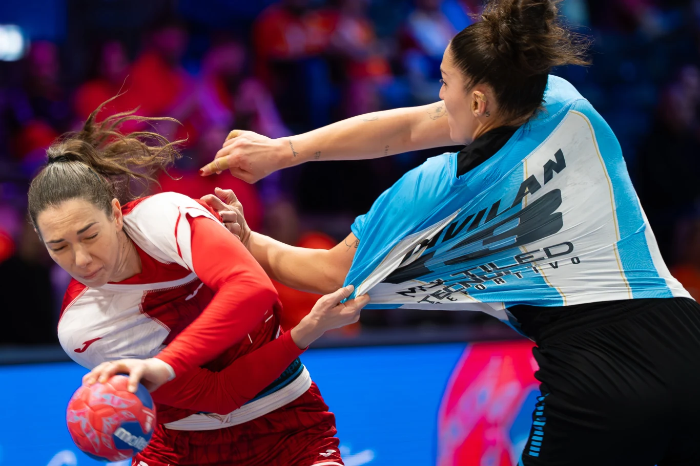 Monika Kobylińska (z lewej) w meczu z Argentyną Dynamiczna walka dwóch zawodniczek podczas meczu piłki ręcznej, jedna z nich chwytając przeciwniczkę za koszulkę, próbuje ją zatrzymać, obie skupione na grze, w tle rozmyte sylwetki kibiców.
