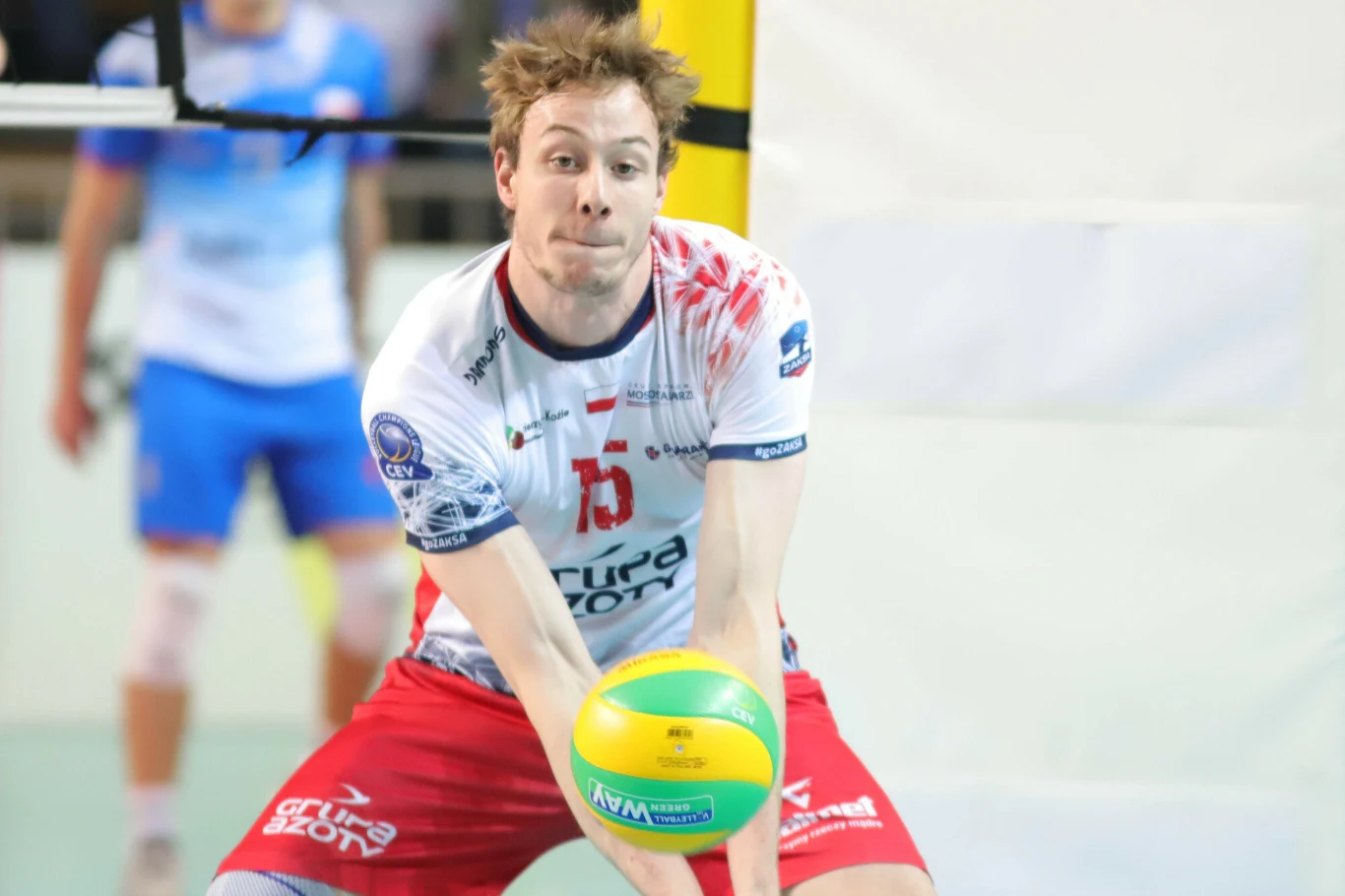 Siatkarz w białej koszulce i czerwonych spodenkach odbija piłkę podczas meczu. W tle znajduje się siatka oraz drugi zawodnik w niebieskim stroju.