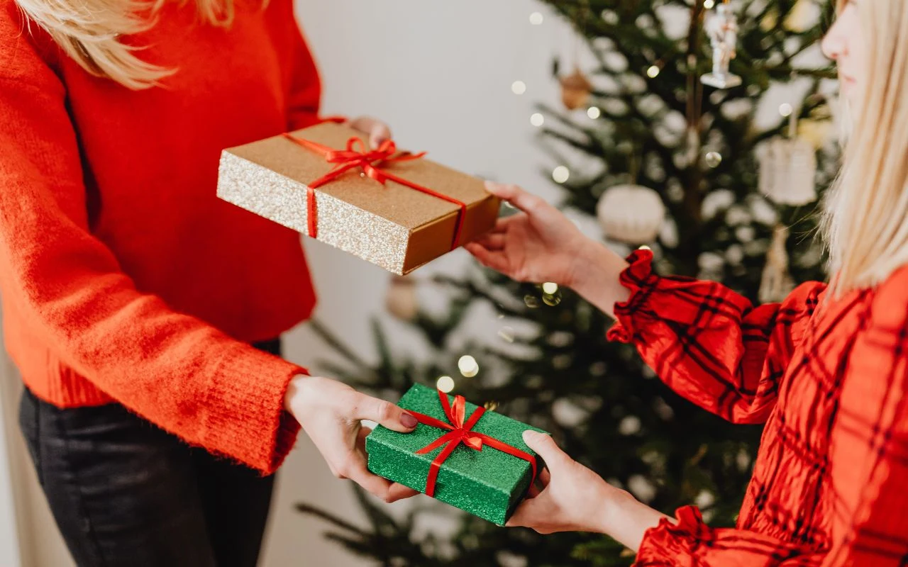 Wymiana prezentów dla niektórych to wielka radość, a dla innych duży stres Dwie osoby wręczają sobie kolorowo zapakowane prezenty, w tle widoczne ozdobione świątecznymi dekoracjami drzewko choinkowe.