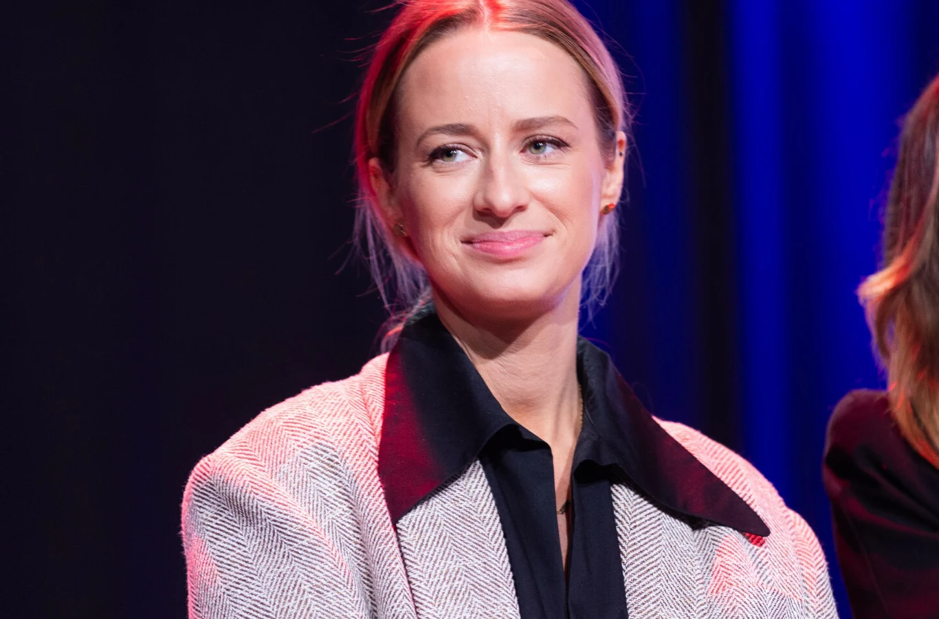 Urszula Dębska Kobieta z jasnymi włosami spiętymi z tyłu, uśmiechnięta, ubrana w jasny żakiet z czarnymi elementami i czarną koszulę, siedzi na tle ciemnego, jednolitego tła oświetlonego światłem w tonacji niebiesko-różowej.