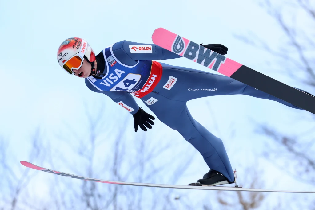 Paweł Wąsek Zawodnik w kombinezonie narciarskim wykonuje skok narciarski w powietrzu, w tle rozmyte gałęzie drzew i jasnoniebieskie niebo.