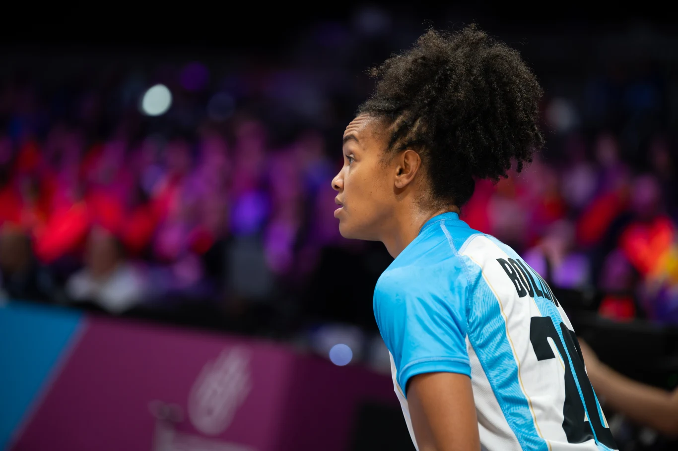 Joana Bolling Zawodniczka siatkówki w sportowym stroju z numerem 10 na plecach, skoncentrowana i obserwująca rozgrywkę na boisku, w tle rozmyta publiczność i kolorowe światła.