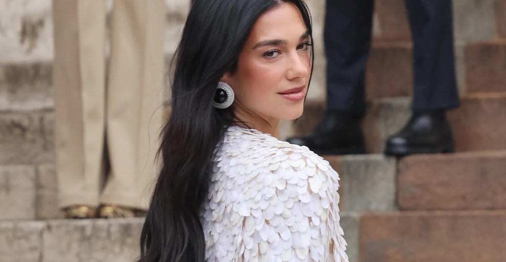Dua Lipa Młoda kobieta z długimi, ciemnymi włosami ubrana w białą kreację z ozdobnymi detalami na ramionach, stojąca bokiem i patrząca przez ramię, w tle schody oraz postacie w eleganckich ubraniach.