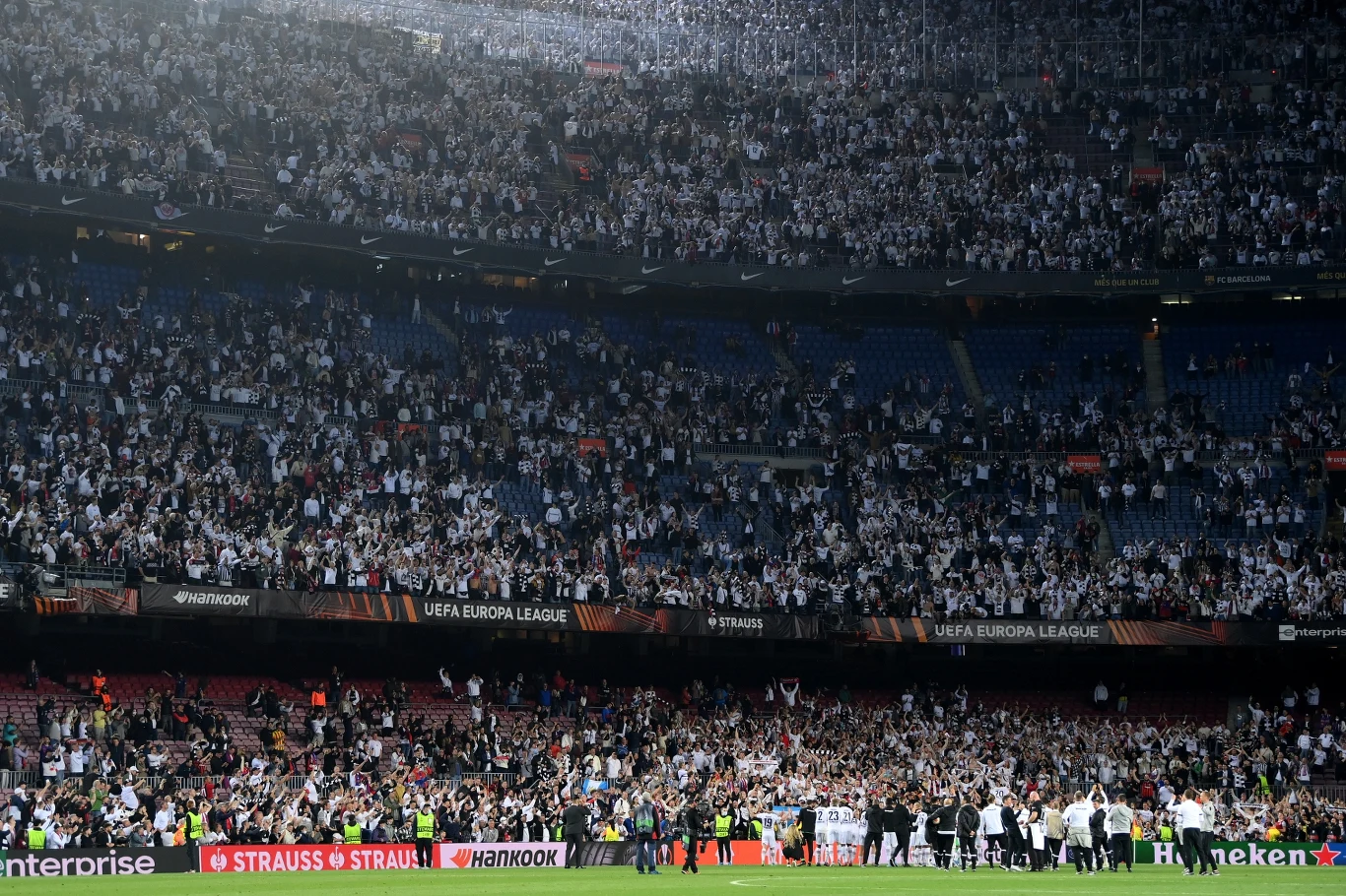 Ogromna liczba kibiców w białych koszulkach zajmuje trybuny stadionu podczas meczu piłki nożnej UEFA Europa League, na murawie widoczna jest grupa osób, prawdopodobnie piłkarzy i sztabu szkoleniowego, wokół banery sponsorów i charakterystyczna architek...