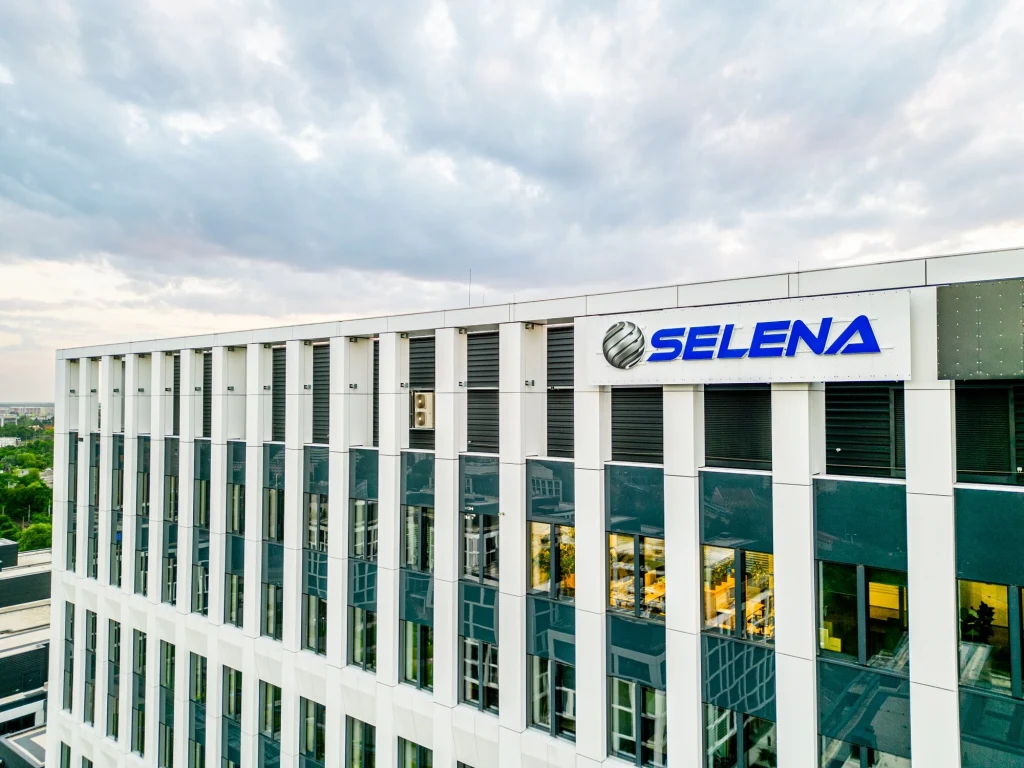 Nowoczesny biurowiec z dużymi oknami, na froncie widoczne logo firmy Selena. W tle pochmurne niebo oraz fragmenty miejskiego krajobrazu.