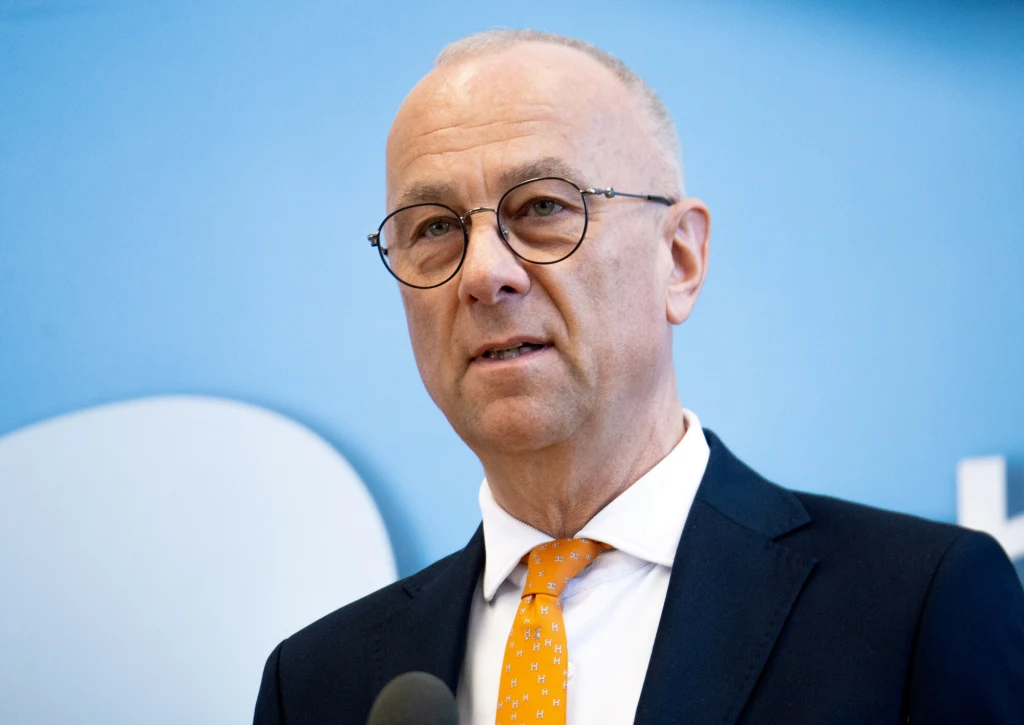 Były marszałek województwa lubuskiego usłyszał wyrok Mężczyzna w okularach i garniturze, z pomarańczowym krawatem, stoi przed niebieskim tłem, patrząc przed siebie podczas przemówienia lub konferencji.