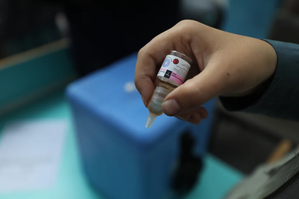 Dłoń trzymająca fiolkę z płynem, prawdopodobnie lekiem lub szczepionką, w tle widoczne nieostre niebieskie pudełko medyczne.