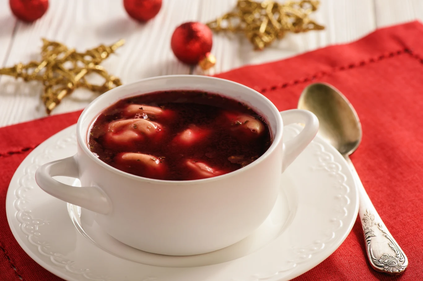 Wigilijny barszcz podaje się najczęściej z domowymi uszkami Biały talerz z barszczem czerwonym i uszkami ustawiony na czerwonej serwetce. W tle złote ozdoby świąteczne oraz czerwone bombki na jasnym stole. Srebrna łyżka leży obok talerza.