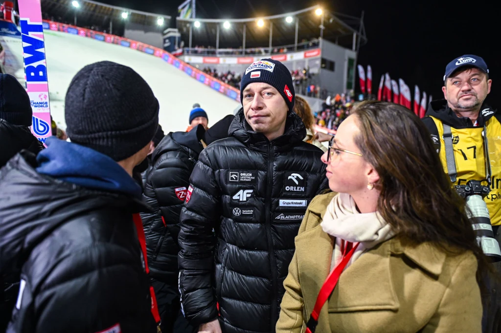 Aleksander Zniszczoł Grupa osób ubranych w zimowe kurtki i czapki rozmawia przy trasie skoków narciarskich podczas zawodów nocą. W tle trybuny, oświetlony stok oraz liczne flagi.