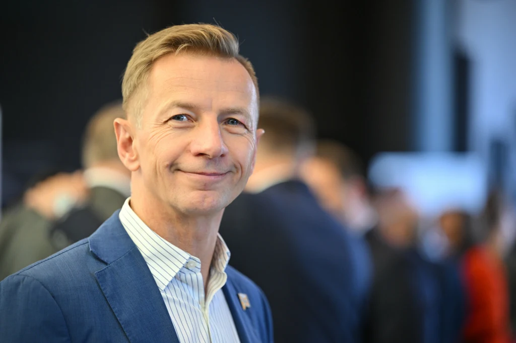 Media: Rafał Kasprzyk kolejnym kandydatem na przewodniczącego Polski 2050 Mężczyzna w średnim wieku w niebieskiej marynarce i jasnej koszuli, uśmiechnięty, patrzy prosto do obiektywu. W tle obecni są inni ludzie, otoczenie wskazuje na wydarzenie formalne lub biznesowe.