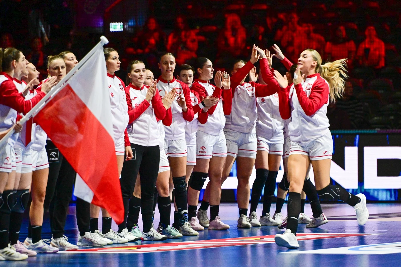 Drużyna kobieca ubrana w czerwono-białe stroje sportowe, stojąca na boisku, jedna zawodniczka biegnie z wyciągniętymi rękami w kierunku koleżanek, z przodu widoczna biało-czerwona flaga Polski.