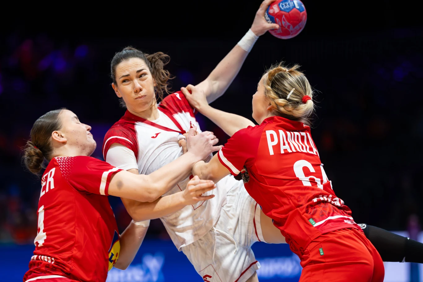 Zawodniczka drużyny w białym stroju próbuje oddać rzut podczas meczu piłki ręcznej, blokowana przez dwie rywalki w czerwonych strojach. Intensywna walka fizyczna oraz skupienie na twarzy zawodniczek podkreślają dynamiczny charakter rozgrywki.