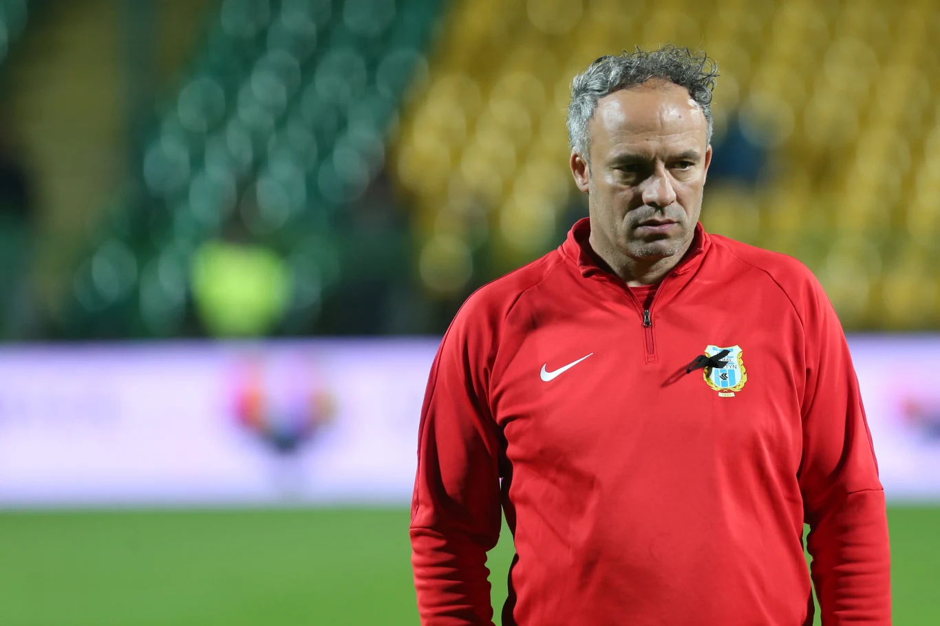 Sylwester Czereszewski Trener w czerwonej bluzie z herbem klubu piłkarskiego stoi na boisku, w tle zamazane trybuny stadionu.