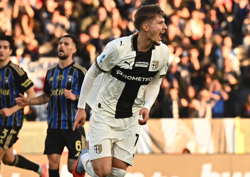 Adrian Benedyczak po zdobyciu debiutanckiego gola w Serie A Piłkarz w białoczarnym stroju z logo Parmy świętuje zdobycie gola na tle widowni, obok inni zawodnicy obserwują sytuację na boisku.