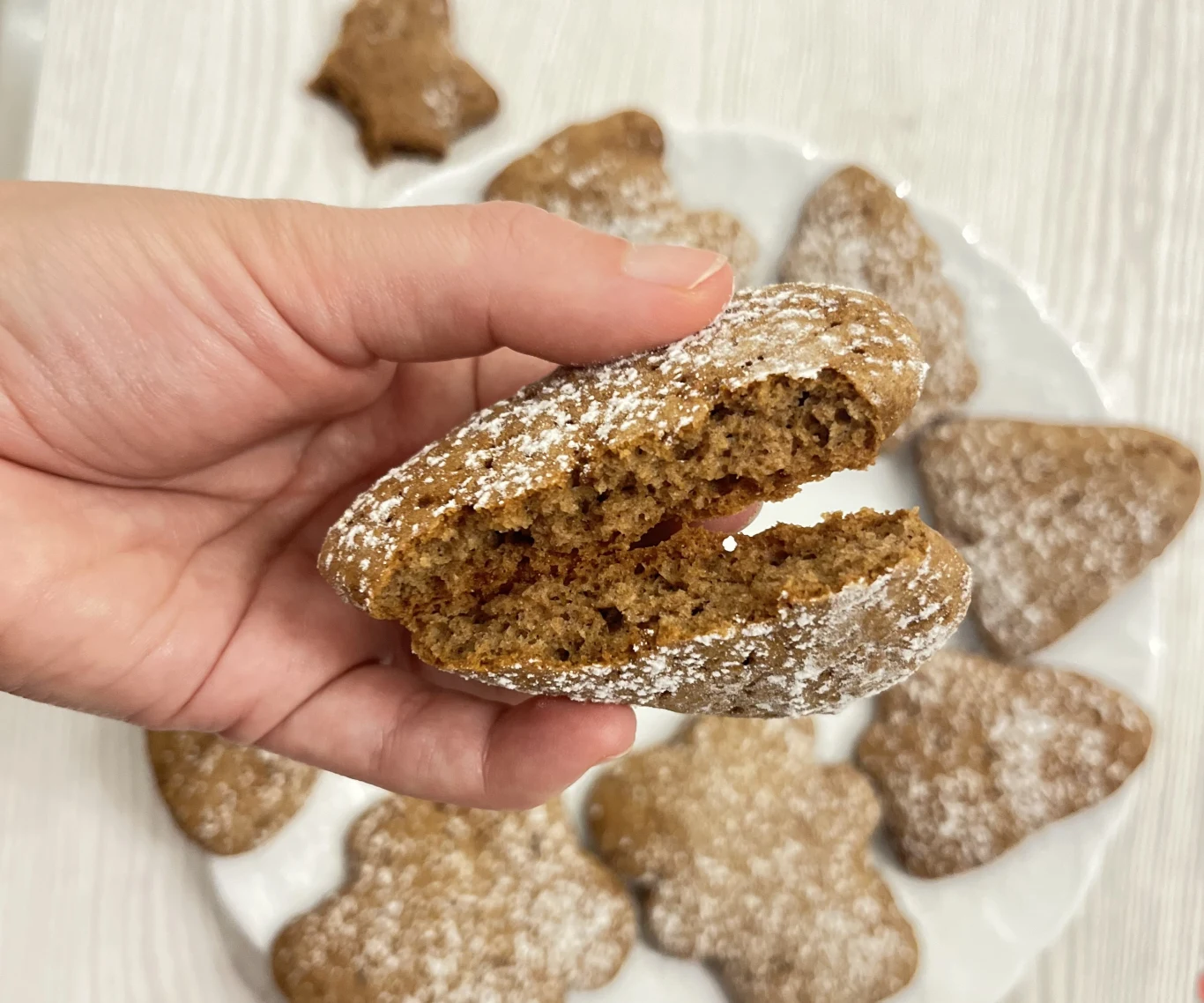 Pierniczki wychodzą puszyste i mięciutkie, a korzenny zapach i smak jest mocno wyczuwalny Ręka trzyma przekrojonego piernika oprószonego cukrem pudrem, na talerzu w tle ułożone są inne pierniki w kształcie serc i gwiazdek.