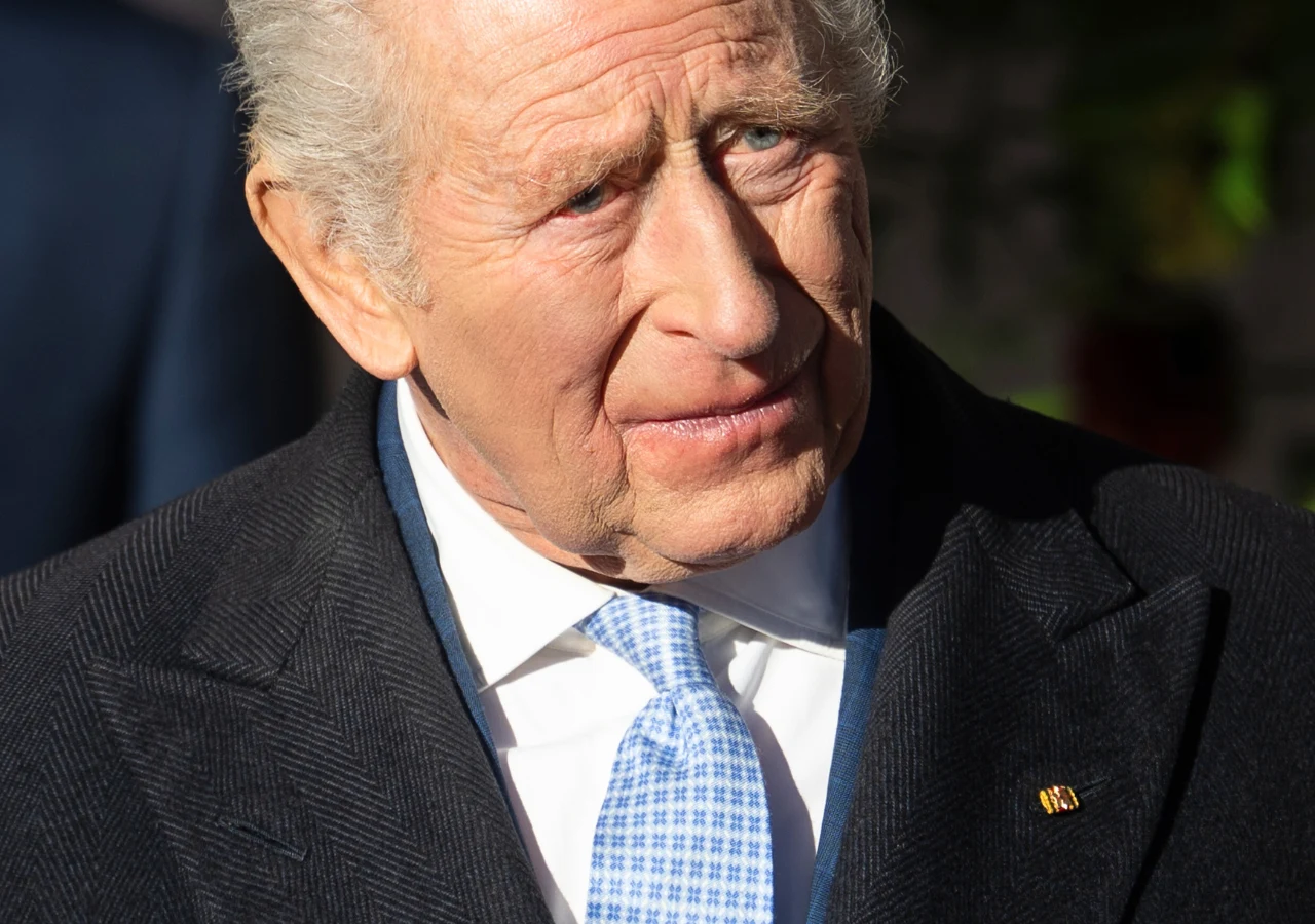 Król Karol III Starszy mężczyzna w eleganckim, ciemnym płaszczu i jasnoniebieskim krawacie, z siwymi włosami i lekko marszczącym się czołem, patrzący lekko w bok w ostrym świetle dziennym.