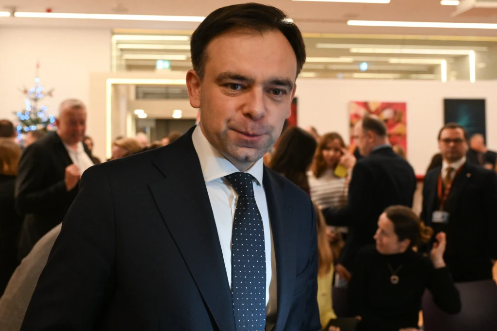 Minister finansów i gospodarki Andrzej Domański chce siedziby Urzędu Unii Europejskiej ds. Celnych w Warszawie Mężczyzna w garniturze i krawacie na tle grupy osób podczas spotkania towarzyskiego lub biznesowego w jasnym, nowoczesnym wnętrzu.