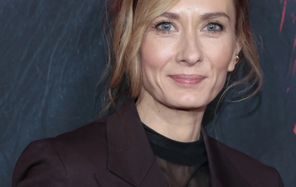 Uśmiechnięta kobieta w eleganckim ciemnym garniturze na tle ciemnej ściany z delikatnym wzorem, włosy częściowo spięte, dyskretny makijaż i spokojny wyraz twarzy.