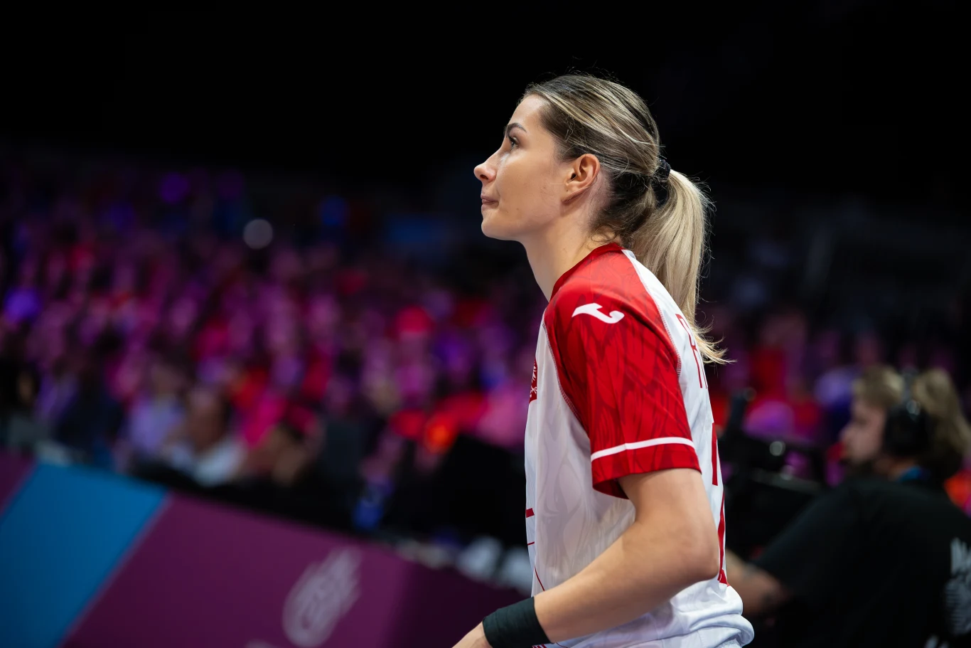 Kobieta w sportowej koszulce z numerem, skoncentrowana, podczas meczu lub rozgrzewki, w otoczeniu widowni i profesjonalnego oświetlenia. Ujęcie wykonane z profilu, w dynamicznym, sportowym otoczeniu.