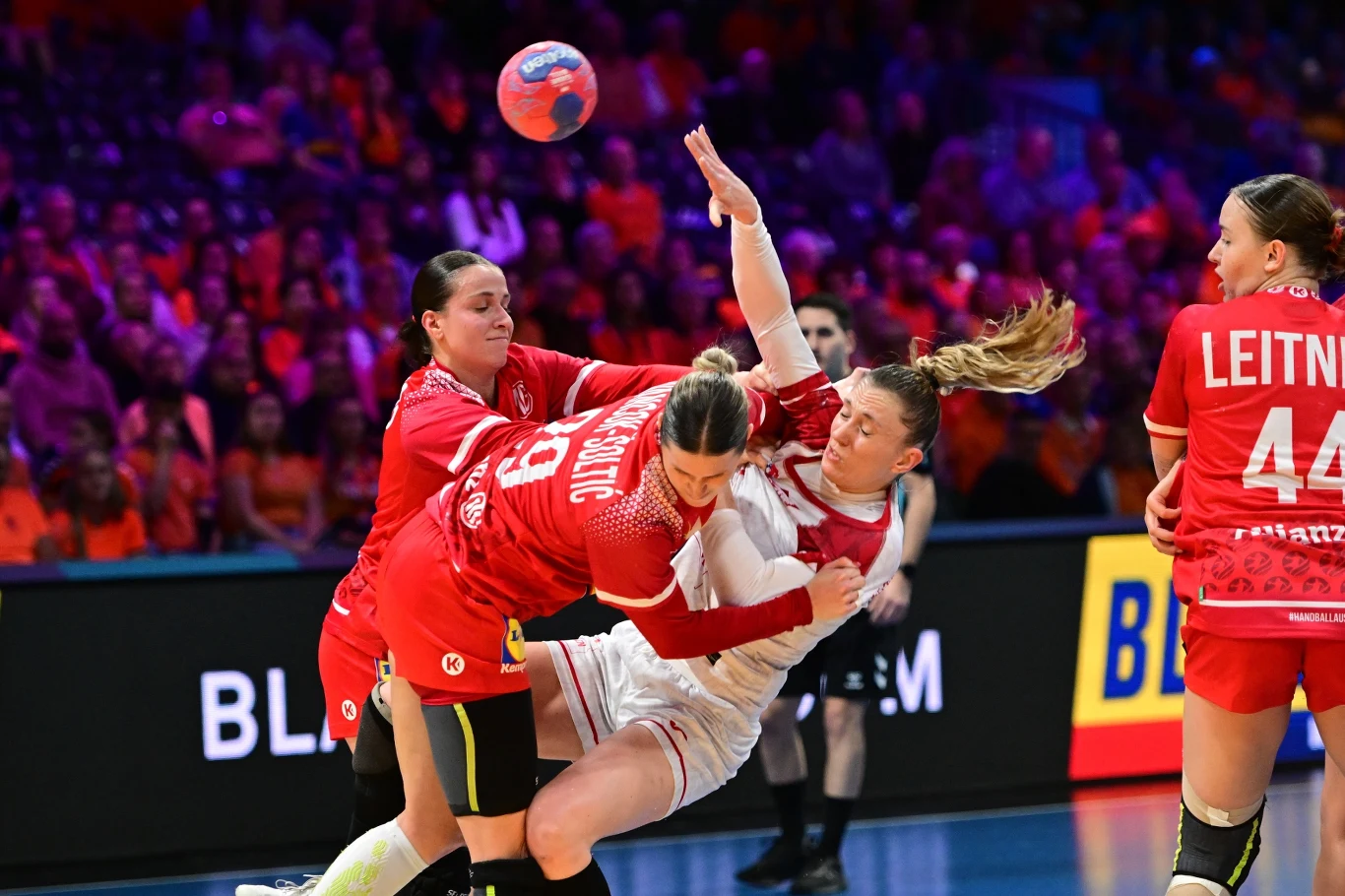 Intensywny moment podczas meczu piłki ręcznej kobiet, zawodniczka w białym stroju jest powstrzymywana przez dwie rywalki w czerwonych koszulkach, piłka znajduje się w powietrzu, a w tle widoczni są kibice i kolorowe oświetlenie hali.