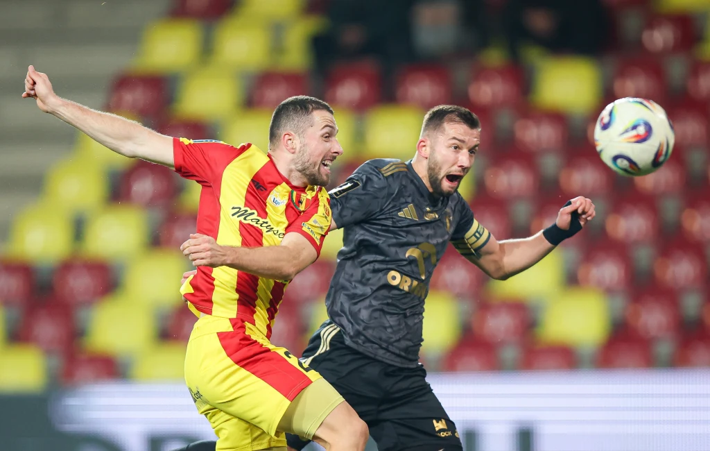 W bramkowej akcji Łukasza Sekulskiego (z prawej) nie zdołał powstrzymać bułgarski defensor Wiktor Popow Dwóch piłkarzy walczących o piłkę w powietrzu podczas meczu, jeden ubrany w żółto-czerwony strój, drugi w czarny, w tle rozmyte kolorowe trybuny stadionu.