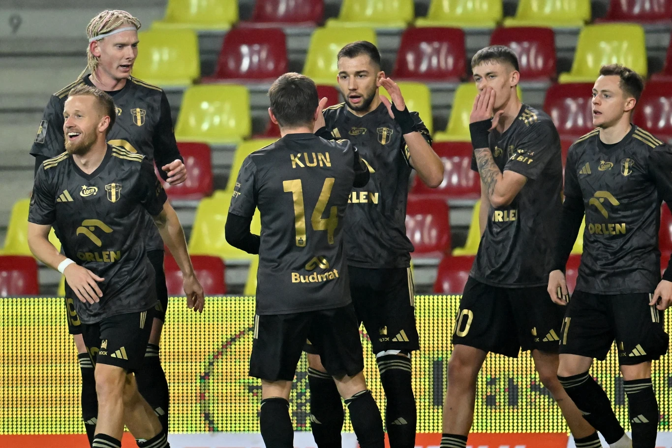 Od 13. minuty płocczanie prowadzili na obiekcie Korony po bramce Łukasza Sekulskiego (w środku) Grupa piłkarzy drużyny w czarnych strojach z nazwiskami i numerami na plecach świętuje na tle pustych, żółto-czerwonych trybun stadionu, zawodnicy wyglądają na zadowolonych i rozmawiają ze sobą.