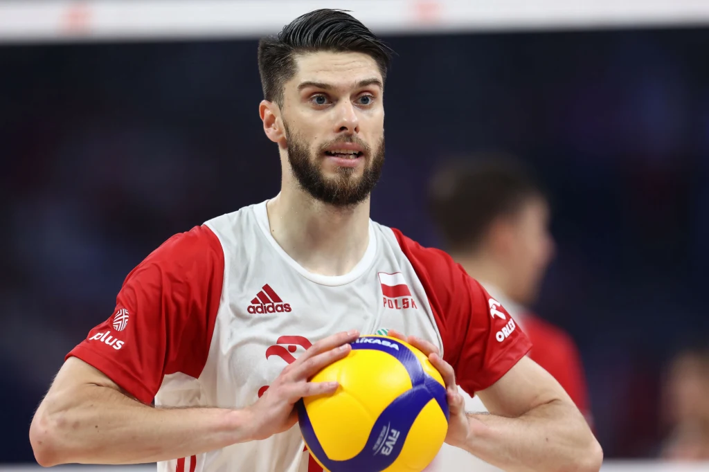 Aleksander Śliwka Siatkarz w stroju reprezentacji Polski trzyma w rękach piłkę do siatkówki, skoncentrowany i gotowy do rozpoczęcia akcji, w tle widoczne rozmyte postaci innych zawodników.