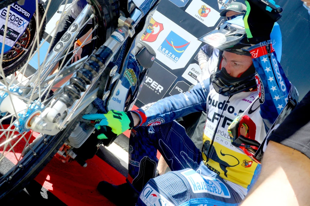 Josh Pickering kuca przed swoim motocyklem Motocyklista w stroju wyścigowym sprawdzający element motocykla żużlowego na tle planszy sponsorów i logotypów.