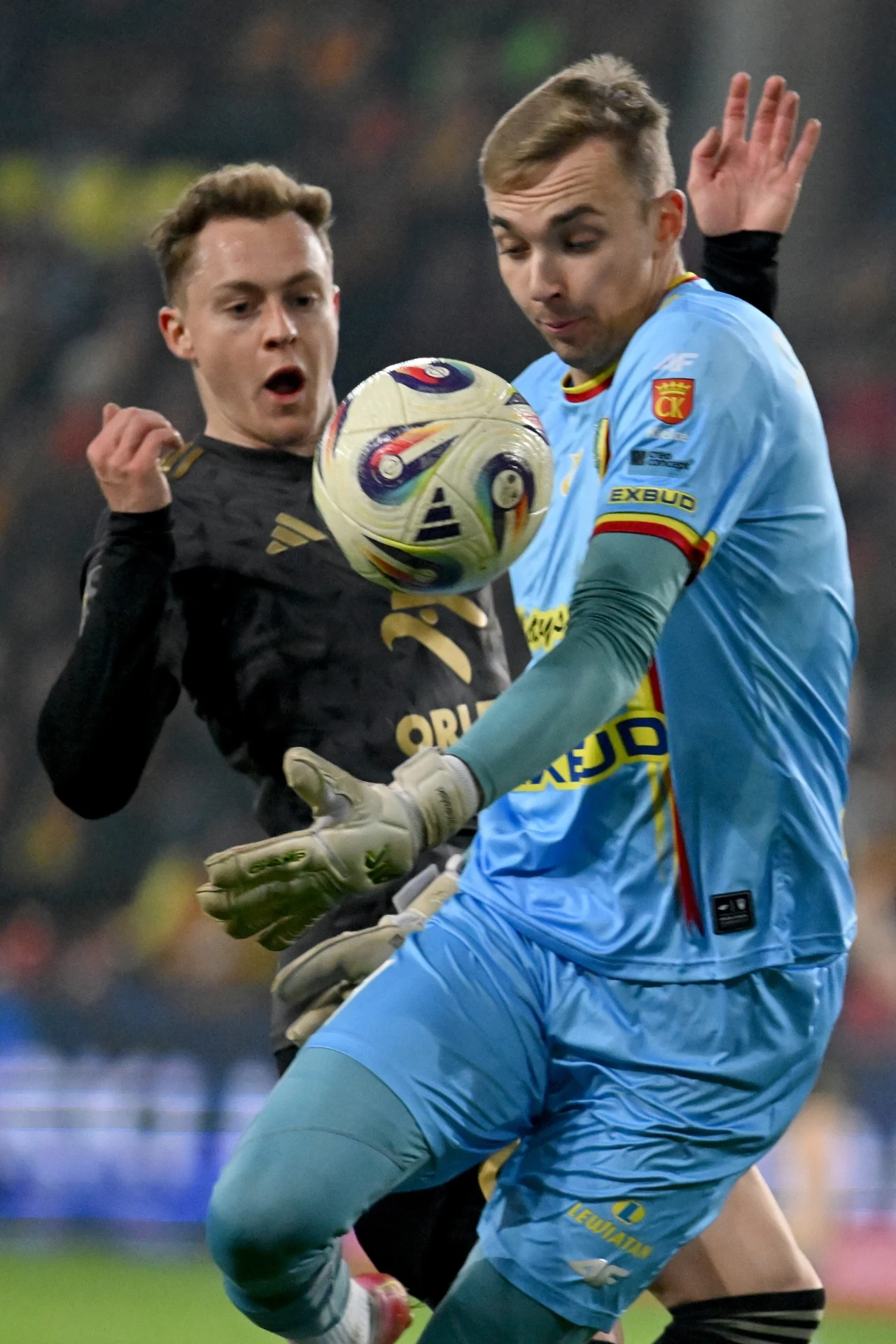 Bramkarz Korony, Xavier Dziekoński, momentami tracił kontrolę sytuacji we własnym polu karnym Dwóch piłkarzy walczy o piłkę, jeden ubrany w czarny strój z logo sponsora, drugi w niebieskim komplecie bramkarskim, skupienie i dynamika gry wyrażone na ich twarzach.