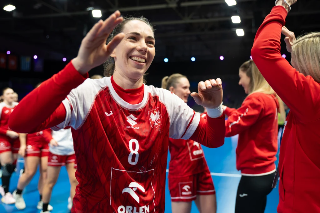 Monika Kobylińska Radość polskich piłkarek ręcznych w czerwonych strojach na parkiecie hali sportowej, uśmiechnięta zawodniczka z numerem 8 na pierwszym planie w otoczeniu koleżanek z drużyny.