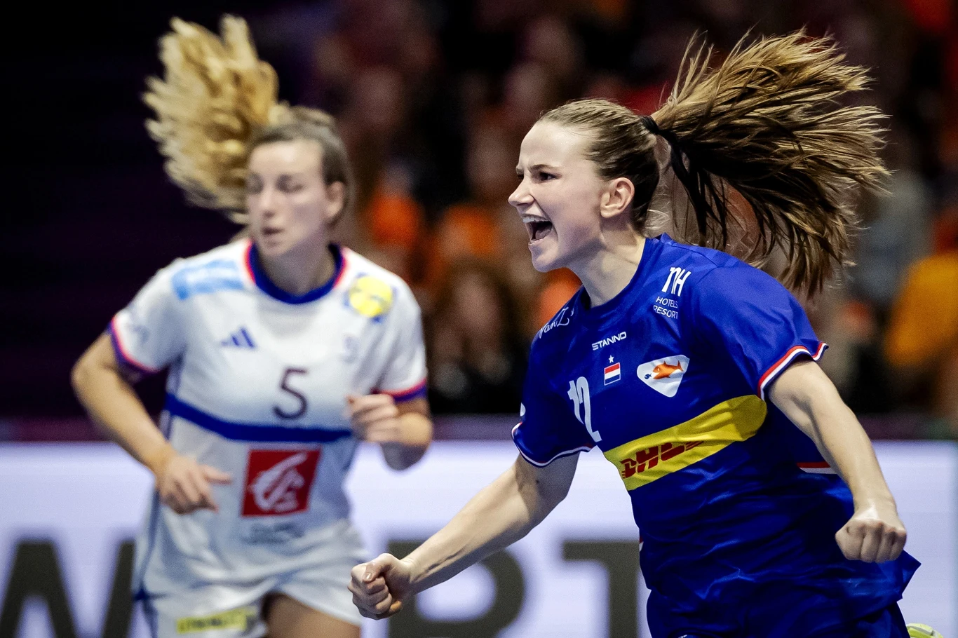 Radość Bo van Wetering, skrzydłowej reprezentacji Holandii Zawodniczka w niebieskim stroju z widoczną radością na twarzy celebruje zdobycie punktu podczas meczu piłki ręcznej, w tle przeciwniczka w białym stroju i rozmyta publiczność.