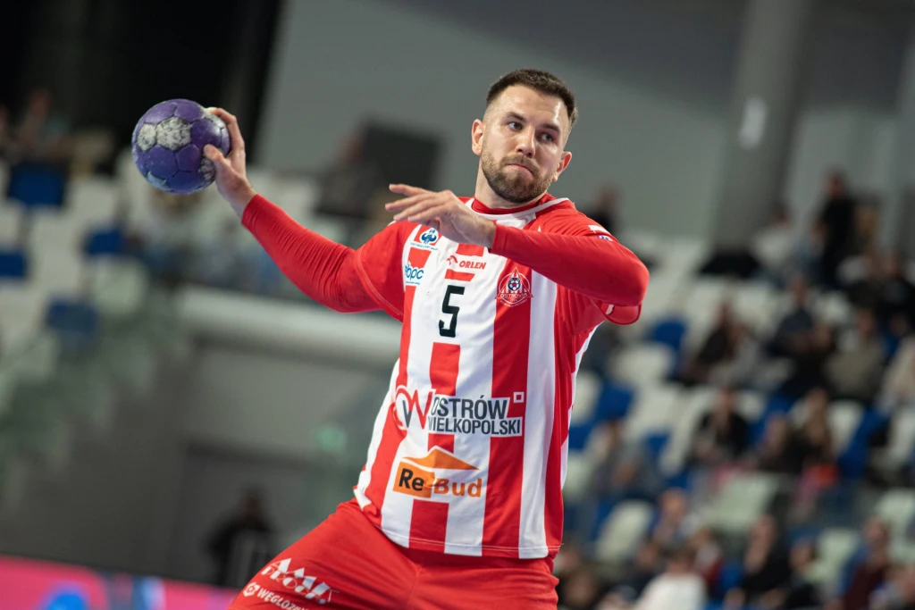 Zawodnik Rebud KPR Ostrovia Ostrów Wielkopolski Zawodnik w stroju sportowym czerwono-białym, z piłką do ręcznej w ręce, przygotowujący się do rzutu podczas meczu, na tle trybun z widownią.