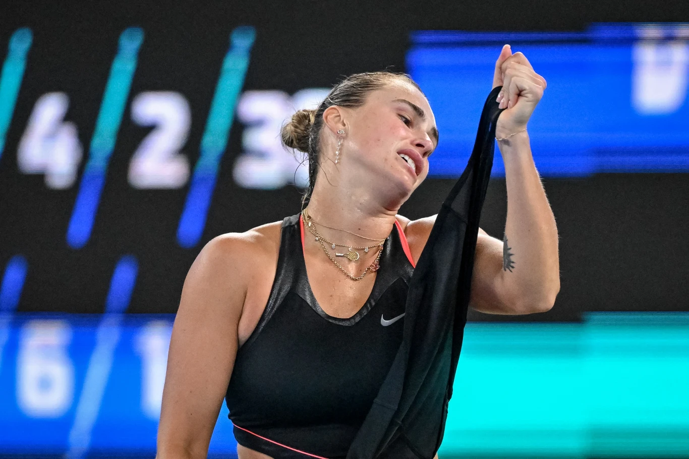 Aryna Sabalenka po raz kolejny zmierzyła się z Naomi Osaką Zawodniczka tenisa ubrana w sportowy top i biżuterię, trzymająca swoją koszulkę, na tle rozmytych cyfr i niebieskozielonego ekranu wskazującego na arenę sportową.