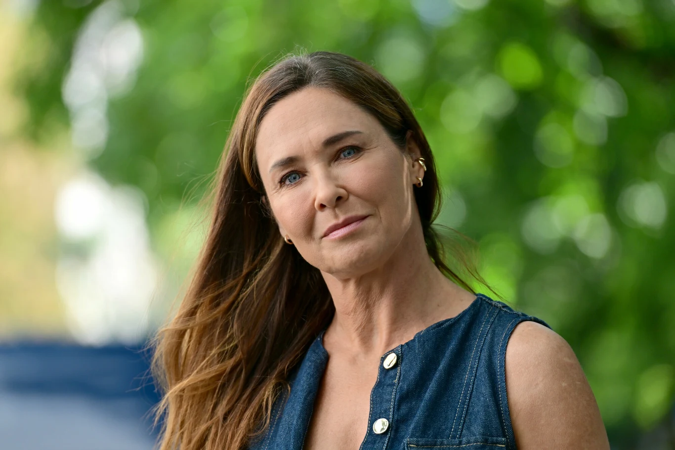 Dorota Naruszewicz Kobieta o długich, brązowych włosach w rozpuszczonych włosach patrzy spokojnie przed siebie, ubrana w ciemnoniebieską koszulę bez rękawów, na tle rozmytej zieleni drzew i lekkiego światła.