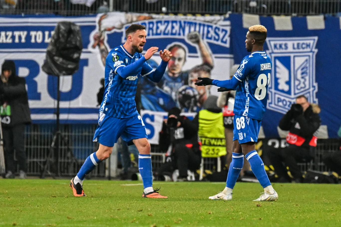 Filip Jagiełło i Taofeek Ismaheel w barwach Lecha Poznań Dwóch piłkarzy drużyny Lech Poznań ubranych w niebieskie stroje celebruje akcję na murawie, w tle widoczna jest grupa kibiców oraz wielki baner klubowy.