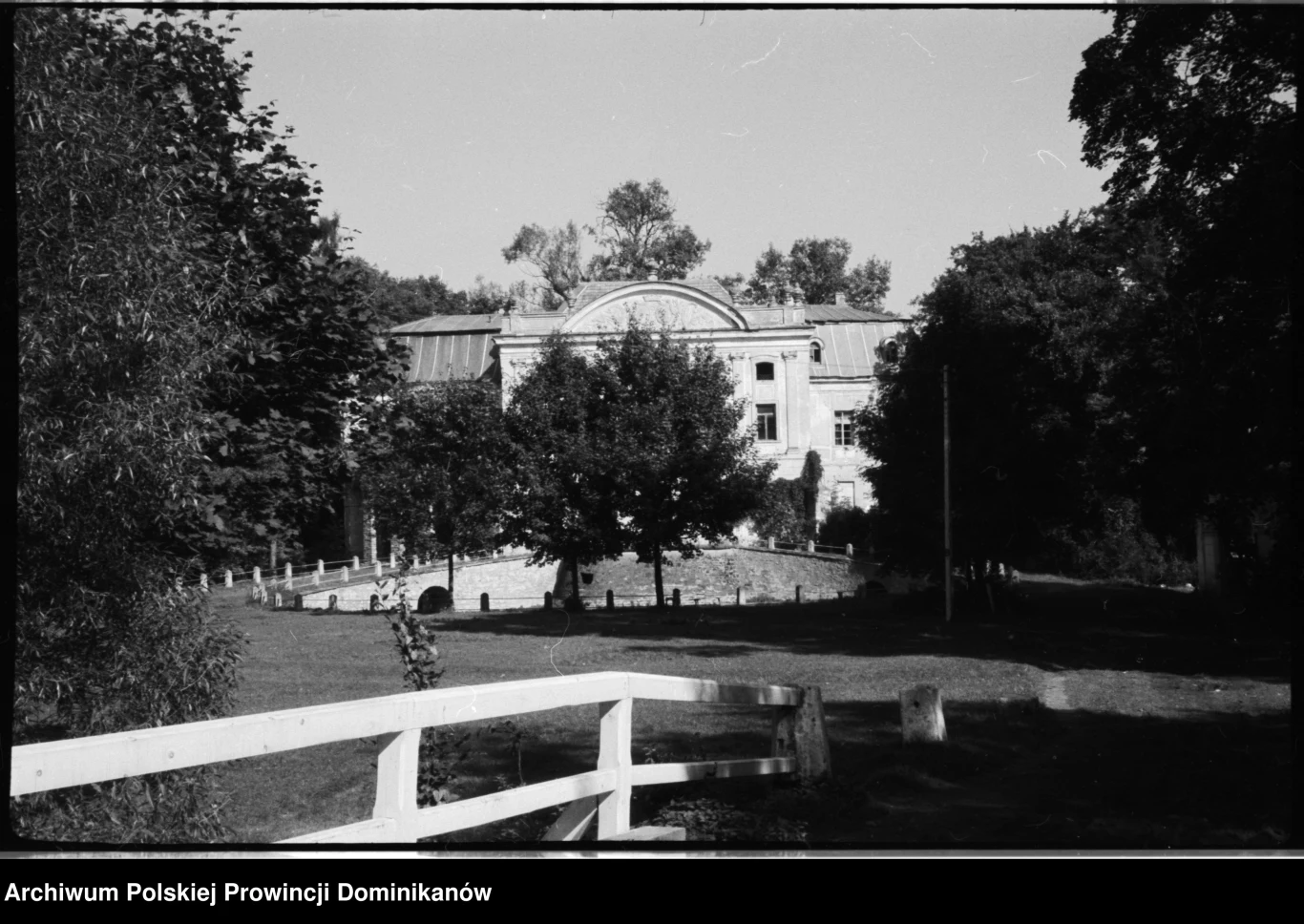 Tak prezentował się pałac w latach 70. XX wieku W centrum zdjęcia znajduje się duży, historyczny budynek otoczony bujną zielenią. Na pierwszym planie widać drewniane ogrodzenie, a za nim rozpościera się przestronny ogród. W tle widoczne są drzewa, a całe otoczenie sprawia wrażenie spokojnego i zadba...