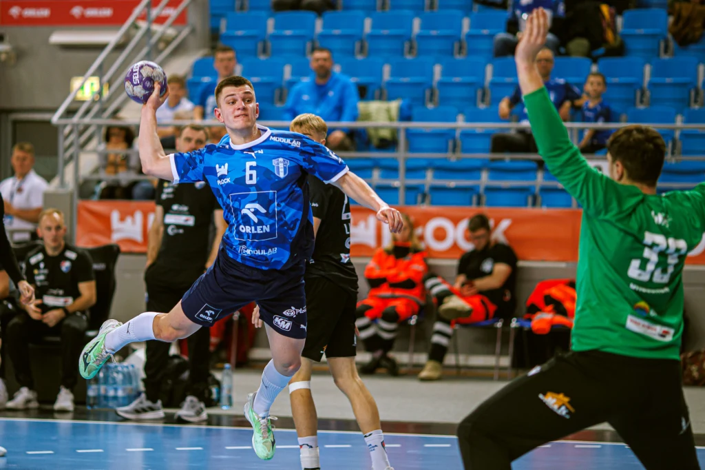 Zawodnik piłki ręcznej w niebieskim stroju przygotowuje się do rzutu na bramkę podczas meczu, a bramkarz w zielonym stroju stara się zablokować strzał, w tle widoczni są kibice i ławka rezerwowych.