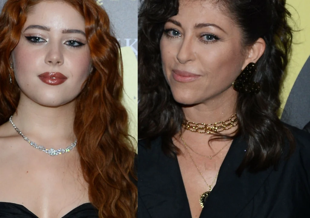 Dwie kobiety w eleganckich strojach, jedna o rudych wydłużonych włosach i wyrazistym makijażu, druga z ciemniejszymi, falowanymi włosami, obie noszą biżuterię i stoją na tle z żółtymi elementami.