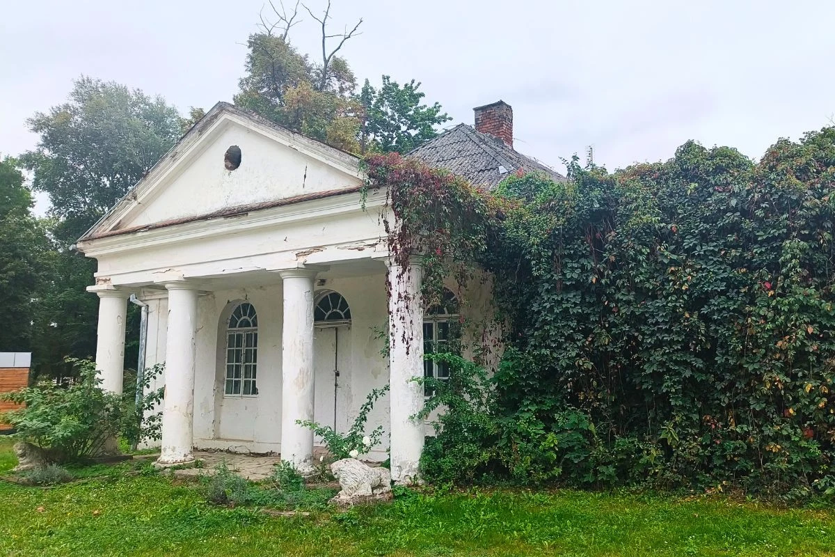 Zobaczysz tu także dawne zabudowania wchodzące w skład całego folwarku Opuszczony dworek z białą fasadą i czterema kolumnami frontowymi, częściowo porośnięty gęstym bluszczem, otoczony zielenią i trawnikiem.