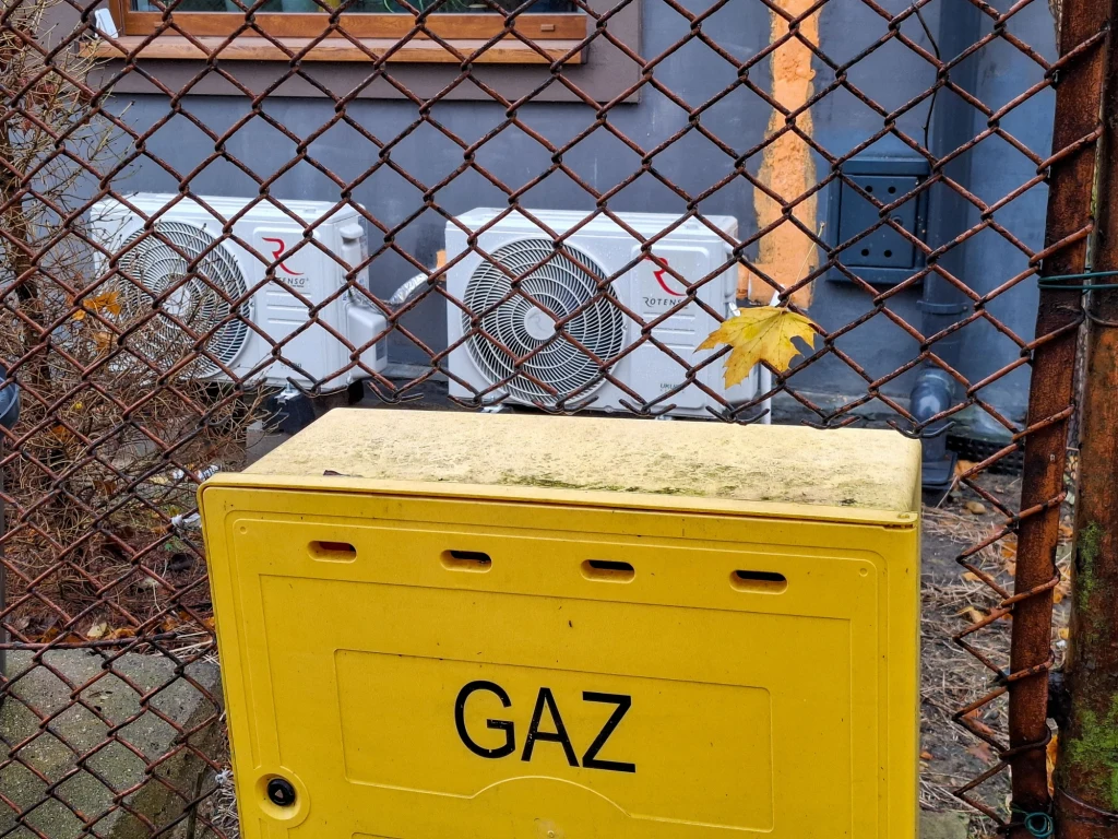 Gaz tylko w połączeniu z OZE? UE zdecydowała. Żółta skrzynka gazowa stoi obok metalowego ogrodzenia z widokiem na dwie białe jednostki klimatyzacyjne zamontowane na ścianie budynku. Nad jednostkami widać fragment okna oraz szarej elewacji budynku.