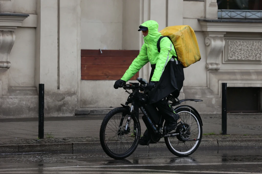 GUS podał dane dotyczące liczby cudzoziemców wykonujących pracę w Polsce Kurier rowerowy w jaskrawozielonej kurtce i z żółtym plecakiem jedzie po mokrej ulicy obok zabytkowego budynku, pada deszcz.