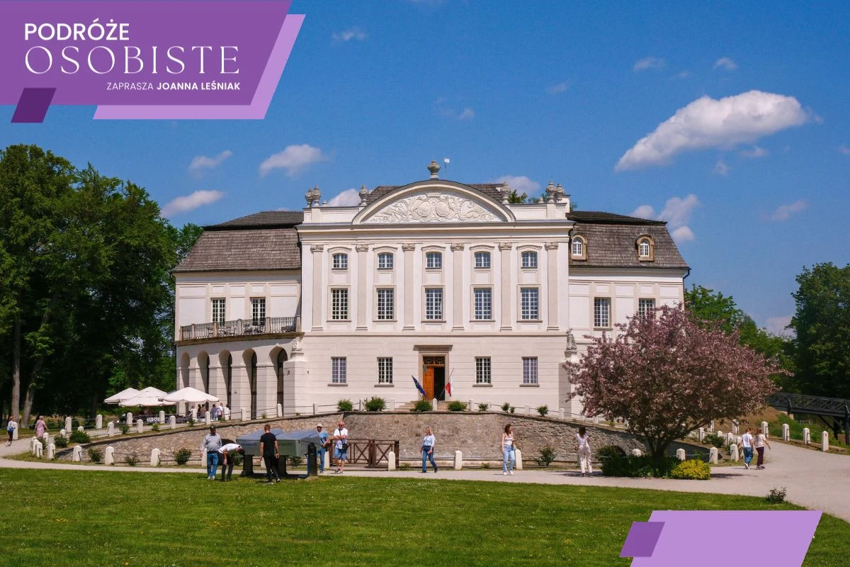 Pałac w Kurozwękach - czy warto go odwiedzić? Elegancki, klasycystyczny pałac otoczony zadbanym ogrodem, przed którym spacerują i rozmawiają niewielkie grupy osób. Po lewej stronie widoczne białe parasole przy stolikach, a po prawej rozkwitnięte drzewo. W tle błękitne niebo z pojedynczymi chmurami.