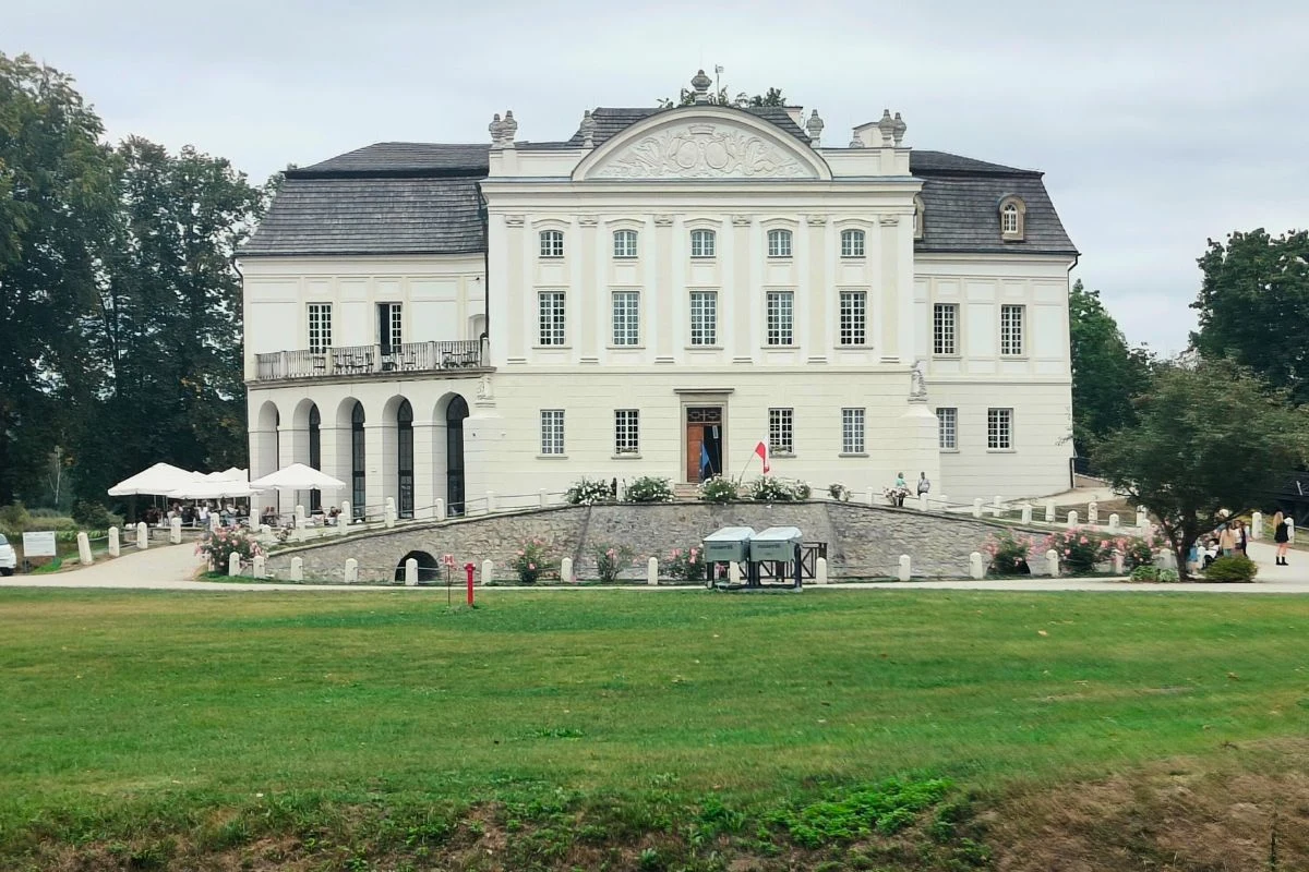 Województwo świętokrzyskie obfituje w ciekawe miejsca. Kurozwęki to jedno z nich Duży biały pałac o klasycystycznej architekturze, stojący na wzniesieniu, z szerokimi schodami prowadzącymi do wejścia głównego. Po lewej stronie taras z parasolami i widocznymi gośćmi, otoczony zielenią i drzewami.