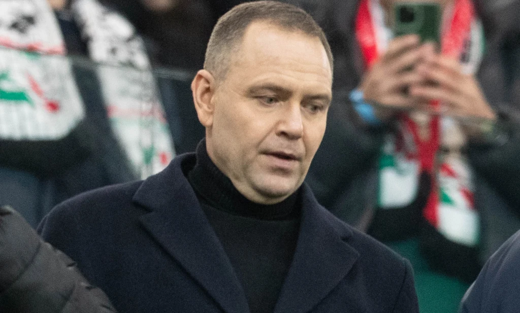 Prezydent Karol Nawrocki na meczu Legia Warszawa - Sparta Praga Mężczyzna w ciemnym płaszczu i golfie znajduje się na pierwszym planie, w tle osoby trzymające telefony oraz szaliki w barwach klubu piłkarskiego.