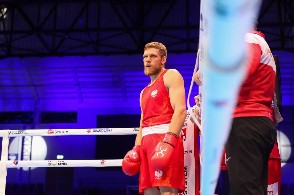 Aleksiej Sewostjanow Bokser w czerwonym stroju z rękawicami znajduje się w ringu, obok niego stoi członek ekipy technicznej w czerwono-białej kurtce. W tle widoczne są barierki oraz niebieskie światła areny.