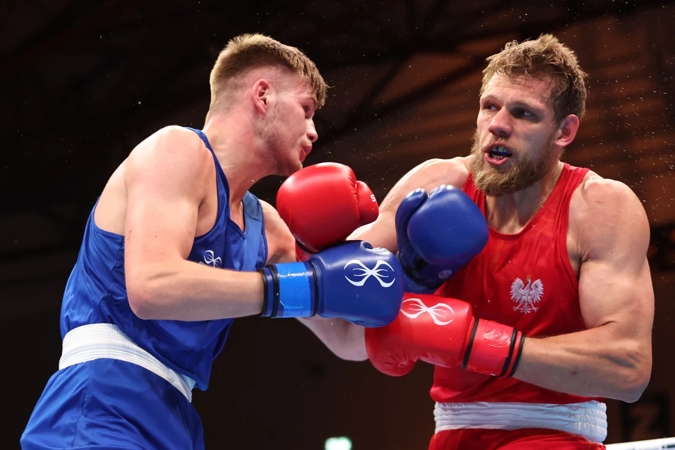 Dwóch bokserów walczy na ringu, ubrani odpowiednio w niebieski i czerwony strój, z widocznym polskim godłem na koszulce zawodnika w czerwonym stroju. Obaj mają podniesione rękawice i są zaangażowani w pojedynek, intensywność walki widoczna na ich twarz...