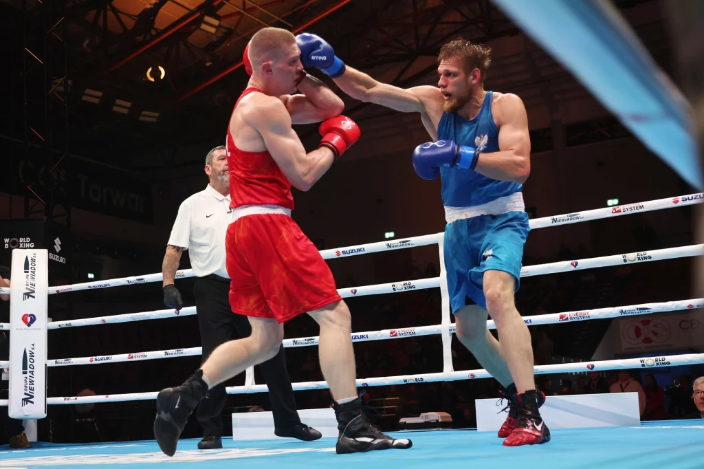 Aleksiej Sewostjanow (z prawej) w finale turnieju im. Feliksa Stamma boksował z Adamem Tutakiem Dwóch bokserów rywalizuje w ringu, jeden ubrany w czerwony strój, drugi w niebieski, w tle widoczny sędzia obserwuje pojedynek, akcja skupia się na dynamicznym wymianie ciosów między zawodnikami.