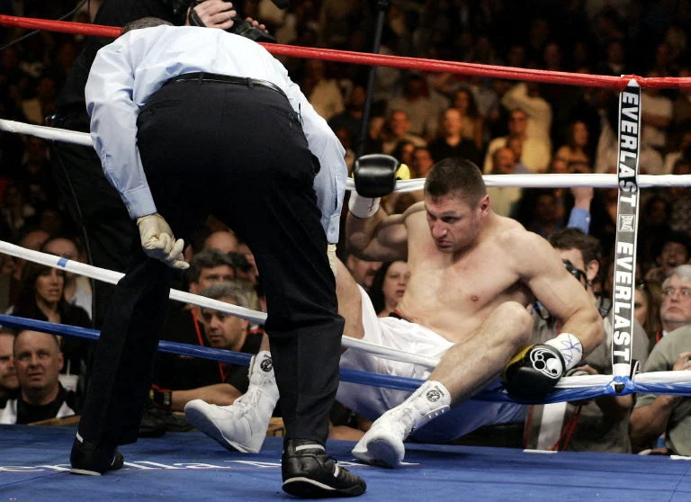 Andrzej Gołota przegrał z Lemonem Brewsterem Bokser w białych spodenkach siedzi na macie ringu bokserskiego, oparty o liny i wyraźnie wyczerpany lub znokautowany. Obok niego sędzia pochyla się, prawdopodobnie odliczając czas. W tle widać tłum kibiców obserwujących przebieg walki.