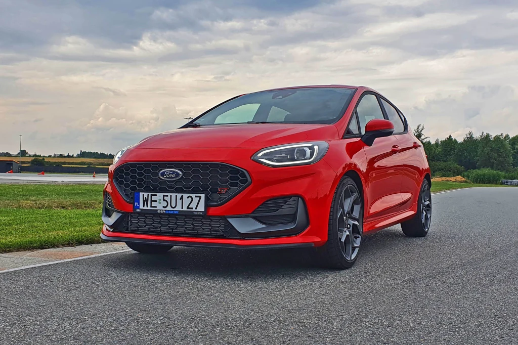 Ford wprowadzi do oferty dwa nowe miejskie elektryki, bazujące na modelach Renault. Bardzo możliwe, że jeden z nich będzie się nazywał Fiesta Czerwony samochód marki Ford Fiesta stoi zaparkowany na asfalcie, w tle widoczna zieleń drzew i pochmurne niebo.